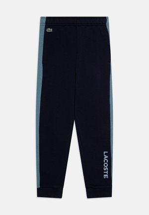Pantalons de survêtement navy avec des panneaux latéraux teal. Doté d'une taille élastique et de poignets côtelés. Logo "LACOSTE" en bleu clair sur le côté gauche.
