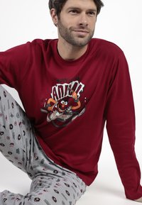 Hombre con una camisa de manga larga roja con un gráfico de un baterista de dibujos animados y pantalones grises con patrones repetidos de personajes de dibujos animados, sentado en el suelo blanco.