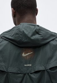 Οπίσθια όψη ενός ατόμου που φοράει ένα σκούρο πράσινο μπουφάν Nike Running με πορτοκαλί στρογγυλό λογότυπο swoosh στην πλάτη κοντά στη κουκούλα.