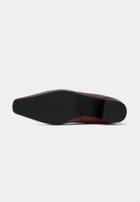 Semelle de chaussure rouge foncé à bout pointu avec un dessous noir texturé et un petit talon bloc, sur un fond blanc uni.
