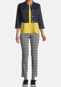 Kvinna klädd i en kort mörk jacka, gul topp, svart-vitrutiga byxor och svarta sneakers, stående mot en enfärgad bakgrund.