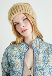 Tan gebreide muts met verticale ribbels. Blauwe met bloemenpatroon gevoerde jas met hoge kraag en grijze piping, gecombineerd met een beige top.