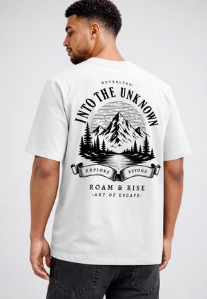 Man wearing white t-shirt with black mountain and tree design and the text "Into The Unknown, Explore Beyond, Roam & Rise, Art of Escape" on the back.