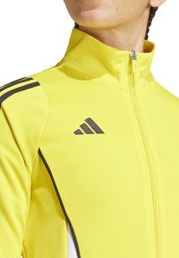 Geel trackjack met rits, voorzien van zwarte strepen op de schouders en een logo op de borst, gemaakt van glad, lichtgewicht materiaal.