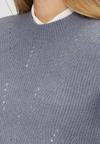 Pull en tricot gris avec un col rond, présentant un motif texturé et des perforations subtiles. Superposé sur une chemise blanche à col.
