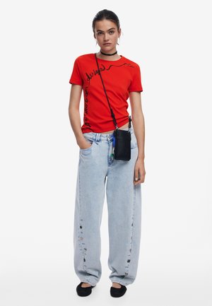 Czerwona bawełniana koszulka z czarnym napisem, zestawiona z jasnoniebieskimi, luźnymi jeansami z guzikami po bokach oraz czarnymi butami wsuwanymi.