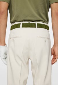 Pantaloni color sabbia con una cintura intrecciata verde oliva; presenta due tasche posteriori con bottoni; guanto da golf bianco parzialmente visibile sulla mano sinistra.