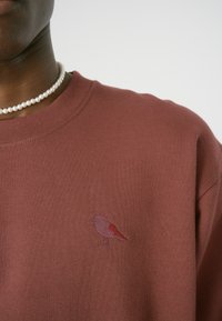 Brauner Sweatshirt mit Rundhalsausschnitt, das auf der linken Brustseite ein kleines gesticktes Vogelmotiv in Pink und Rot aufweist.