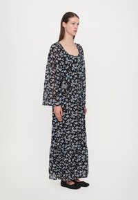 Abito maxi con un motivo floreale nero, maniche lunghe, scollatura arricciata e orlo a strati. Accentuato con una cintura nera e scarpe flat.
