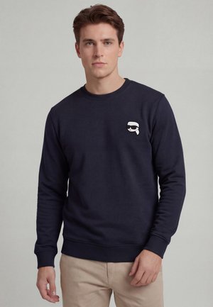 Young man with light skin and brown hair wearing a navy sweatshirt with a small white cartoon logo on left chest and beige pants.