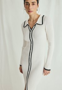 Ribbed knit dress in white with black trim, button front, and collar detail. Features long sleeves and a side slit.