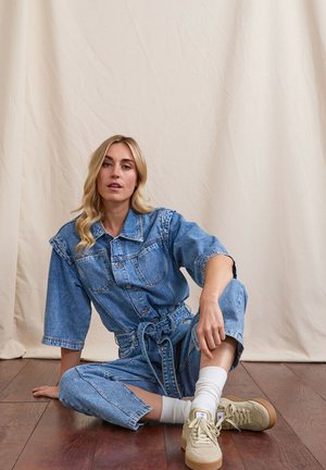 Femme blonde assise sur un sol en bois, portant une combinaison en denim ceinturée, des chaussettes blanches et des baskets beiges, devant un fond beige uni.
