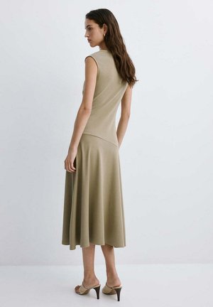 Mujer con cabello largo que lleva un vestido midi beige sin mangas y sandalias beige de tacón alto, de pie frente a un fondo claro y liso.