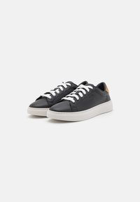 Sorte lærresk sneakers med hvite snører og et teksturert tab på hælen. Har en flat hvit såle og minimalistisk design.