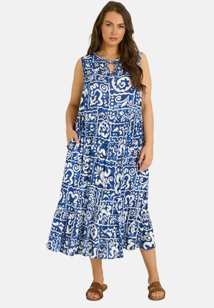 Vrouw draagt een mouwloze midi-jurk met blauw-wit patroon en een gerimpelde zoom, en bruine instapsandalen, staat met handen in de zakken.