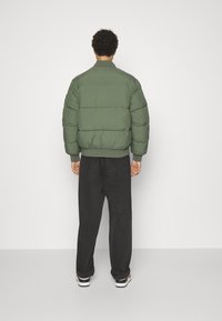 Groene pufferjas met een opstaande kraag en horizontale naden, gecombineerd met zwarte losse broek. De jas heeft elastische manchetten en zoom.