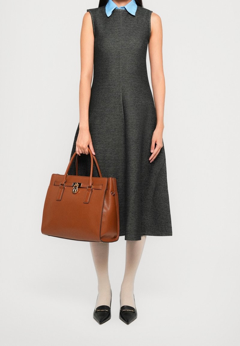 Femme portant une robe sans manches gris foncé avec un col bleu, tenant un grand sac à main en cuir marron, des collants beige et des chaussures noires pointues.