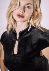 Sort stoftop med høj krave, nøglehulåbning og korte ærmer. Blød tekstur med en let glans; har dekoreret knudeknaplukning.