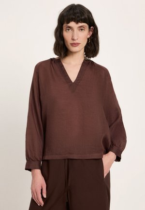 Frau mit kurzen dunklen Haaren, die eine lockere braune Bluse mit langen Ärmeln und V-Ausschnitt sowie passende braune Hose trägt, vor einem schlichten Hintergrund stehend.