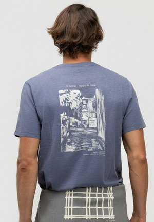 Hombre con cabello de longitud media lleva una camiseta azul con un estampado abstracto en blanco de una escena callejera y texto en la espalda, y una prenda de cuadros gris en la cintura.