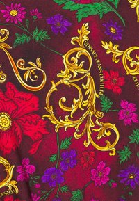 Flerfärgat tyg med djärva blommönster i rött, rosa, lila och grönt, med intrikata guldfärgade barockdetaljer och varumärkes text.