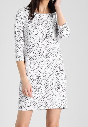 Femme portant une robe blanche mi-longue aux manches trois-quarts avec un motif abstrait noir à points.