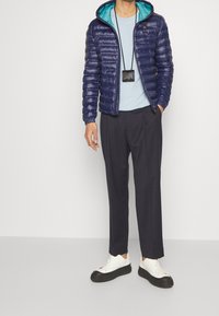 Giubbotto puffer blu navy con fodera teal, chiusura con zip e polsini elasticizzati. Abbinato a una t-shirt azzurro chiaro, pantaloni scuri e sneakers bianche.