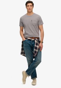 Grijze katoenen t-shirt met zak, blauwe denim jeans, geruite blouse om de taille geknoopt, beige hoge sneakers. Eenvoudig, casual ontwerp.