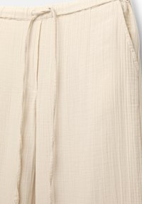Beige vida byxorna gjorda av texturerad tyg, med ett elastiskt midjeband med dragsko och sidofickor.