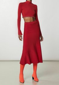 Červený dvoudílný outfit s krátkým dlouhým rukávem a řetízkovými detaily, doplněný rozevlátou midi sukní. Stylizováno s oranžovými kozačkami po stehna.