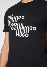 T-shirt noir avec texte blanc déformé et répété "HUGO" sur la poitrine, porté par une personne visible du cou à la taille.