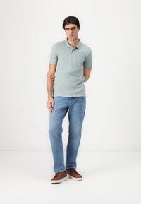 Lys grønn polo skjorte laget av bomull, kombinert med blå denim jeans. Modellen har på seg brune joggesko og står mot en hvit bakgrunn.