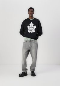 Hanorac negru cu logo-ul alb "Toronto Maple Leafs" și grafică cu frunză. Purtați peste o cămașă de culoare verde deschis, asortat cu blugi gri și pantofi negri.