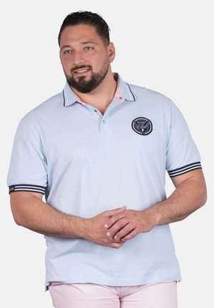 Homme barbu portant un polo de rugby bleu clair avec des poignets de manches à rayures noires et un logo, debout les mains jointes.