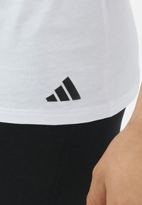 Weißes T-Shirt aus Baumwolle mit einem schwarzen Adidas-Logo in einem dreieckigen Design auf der unteren linken Seite, kombiniert mit schwarzen Hosen.