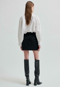 Žena s dlouhými hnědými vlasy, oblečená v bílé blůze s nabíranými rukávy, černé mini sukni a černých vysokých kozačkách na podpatku, stojí s pohledem odvráceným.