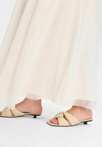 Beige slipper sandalen met een geknoopt ontwerp, met een gladde textuur en een lage zwarte hak, gecombineerd met een lichte, vloeiende crème rok.