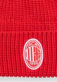 Bonnet rouge tricoté avec une texture côtelée, arborant un emblème ovale blanc affichant "ACM", une croix, et "1899" à l'avant.