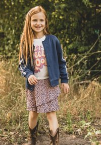 Blauwe jas met witte strepen, wit T-shirt met afbeelding, rok met bloemenpatroon en ruches, en bruine cowboylaarzen. Natuurlijke achtergrond.