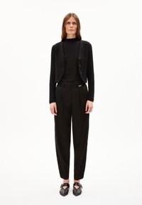 Black knit cardigan with buttons over a black ribbed top, paired with tailored black trousers and flat black shoes with ankle straps.