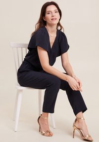 Tuta blu navy con scollatura a V e maniche corte, caratterizzata da una vestibilità rilassata. Abbinata a sandali con tacco oro e semplici dettagli gioiello.
