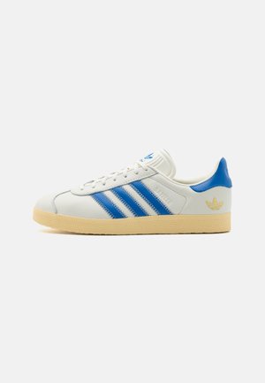 Baskets basses blanches avec des bandes bleues, languette de talon bleue, semelle beige et logo trefoil Adidas doré sur le côté.