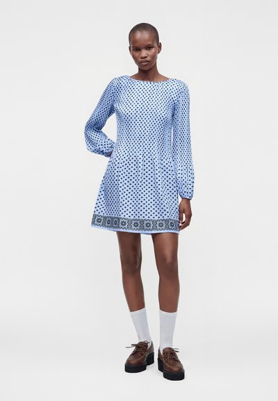 Femme aux cheveux courts portant une robe bleue à manches longues à motifs, des chaussettes blanches et des chaussures à plateforme marron, debout avec une main sur la hanche.