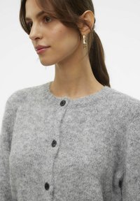 Cardigan en laine grise avec un col rond, texture douce et trois boutons noirs sur le devant. Coupe légèrement oversize et apparence confortable.