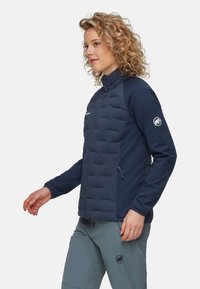 Marinblå puffjacka med blixtlås framtill, horisontell quiltad design på kroppen, ärmar i stretchmaterial och sidofickor med blixtlås.