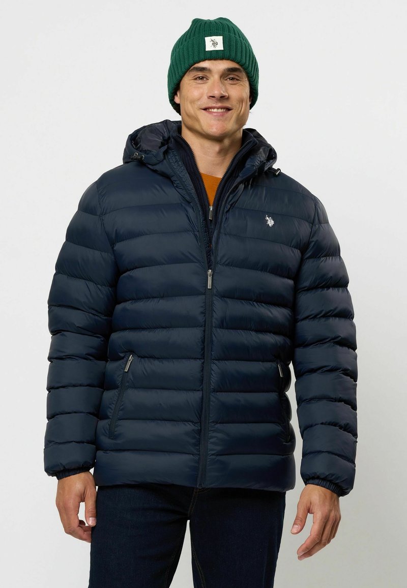 Jūras spēku pufers jaka ar kapuci, ar quilted dizainu, priekšējo rāvējslēdzēju un sānu kabatām. Apvienota ar zaļu ribota beanie cepuri un teksturētu apkakli.