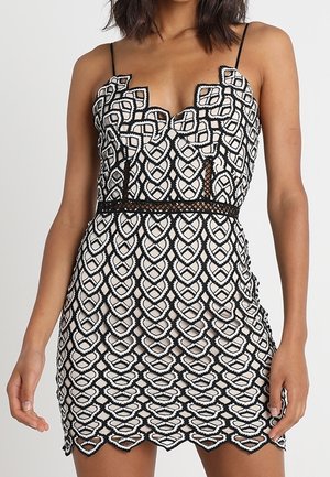 Svart och vit klänning med en vågformad nederdel, intrikat geometrisk spetsöverdrag, justerbara spaghettiband och en figurnära silhuett.