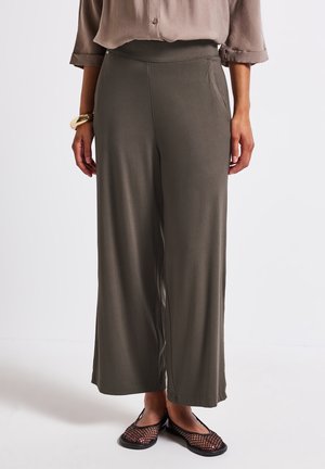 Nainen, jolla on yllään väljät oliivinvihreät leveälahkeiset housut, taupenvärinen, napitettu paita käännetyillä hihoilla, mustat verkkomateriaalista tehdyt liukusandaalit ja kultainen rannekoru.