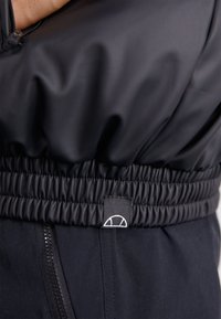 Chaqueta acolchada negra con cintura fruncida, textura suave y un parche de logo. Cierre con cremallera y costuras visibles.
