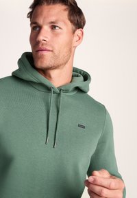 Green hooded sweatshirt made of soft, textured fabric. Features a drawstring hood and small logo on the chest. Simple, casual design.